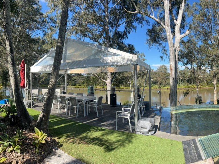 Clear-roof gazebo over a dining table and white chairs beside a backyard pool and river lawn